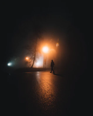 Atmospheric photograph of a lone figure walking through a foggy, dimly lit street.