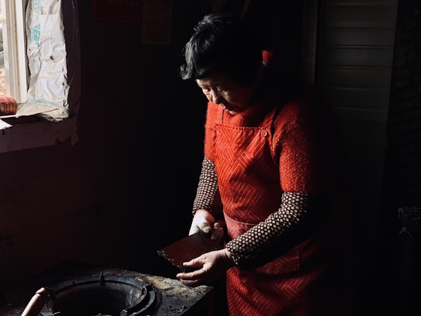 An older woman is standing in a dimly lit room, focusing intently on a task in front of her. She is wearing a red sweater with a patterned apron over it. The light from a nearby window casts a warm glow, highlighting her face and hands as she works. The space appears to be a kitchen with a stovetop visible at the edge of the image.