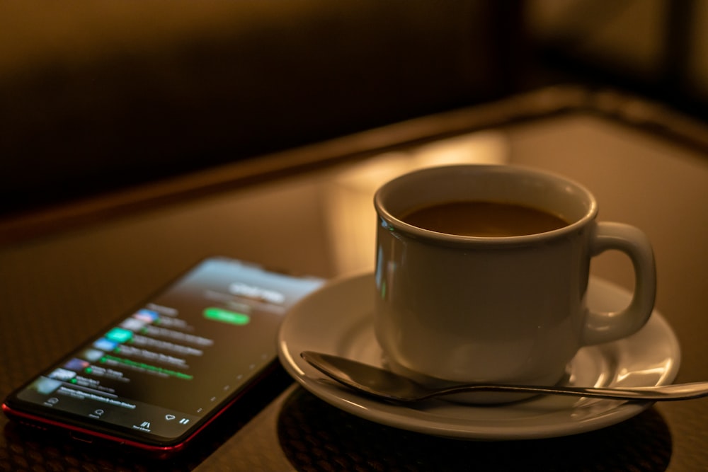 white ceramic cup on saucer beside black smartphone