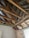 Exposed ceiling beams and wooden rafters in a room under renovation. The room has one white-painted wall and a partially brick wall next to a window. The structure of the ceiling shows signs of deterioration and is partially open to the outside.