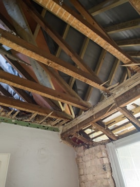 Exposed ceiling beams and wooden rafters in a room under renovation. The room has one white-painted wall and a partially brick wall next to a window. The structure of the ceiling shows signs of deterioration and is partially open to the outside.
