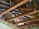 Exposed attic space with wooden beams crossing in multiple directions. There is visible insulation material and some debris. Metal storage containers and cardboard boxes are stored on top of the beams. The wooden structure of the roof is visible, with the underside of roof tiles in sight.
