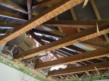 Exposed attic space with wooden beams crossing in multiple directions. There is visible insulation material and some debris. Metal storage containers and cardboard boxes are stored on top of the beams. The wooden structure of the roof is visible, with the underside of roof tiles in sight.
