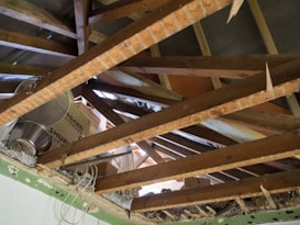 Exposed attic space with wooden beams crossing in multiple directions. There is visible insulation material and some debris. Metal storage containers and cardboard boxes are stored on top of the beams. The wooden structure of the roof is visible, with the underside of roof tiles in sight.