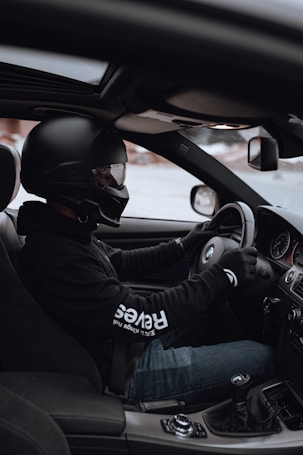 A person wearing a black helmet and gloves is driving a car with a manual transmission. They are seated in the driver's seat, holding the steering wheel, and the gear stick is visible. The interior of the car is modern and includes visible features such as the dashboard and rearview mirror.