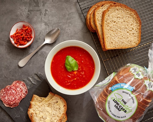 A bowl of vibrant tomato soup garnished with a basil leaf is placed on a gray surface. Surrounding the bowl are slices of fresh bread, both plain and buttered, along with a small dish of chopped red peppers. A few slices of salami are arranged artfully next to the buttered bread, and a spoon lies beside the red peppers. A package of low-carb bread is visible, emphasizing a focus on healthy eating.