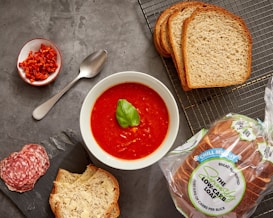 A bowl of vibrant tomato soup garnished with a basil leaf is placed on a gray surface. Surrounding the bowl are slices of fresh bread, both plain and buttered, along with a small dish of chopped red peppers. A few slices of salami are arranged artfully next to the buttered bread, and a spoon lies beside the red peppers. A package of low-carb bread is visible, emphasizing a focus on healthy eating.