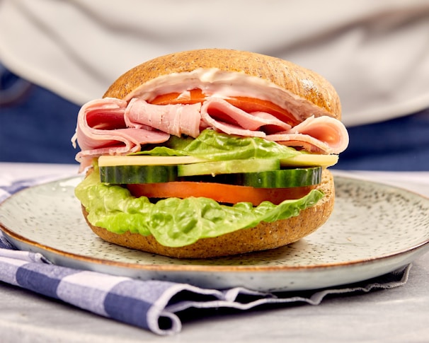 A close-up view of a sandwich featuring slices of ham, layers of fresh lettuce, cucumber, tomato, and cheese, all placed between halves of a whole grain bun. The sandwich is presented on a speckled plate, atop a blue and white checkered napkin.