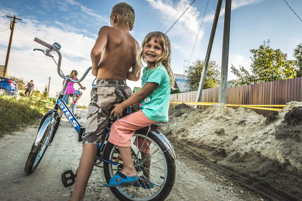 Two children laughing together as they balance on their vivid green Tinywheelz bikes near a leafy playground.