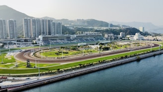 An expansive horse racing track set against a backdrop of high-rise buildings and lush green hills. The area is well-organized with clear visibility of different lanes and seating arrangements. The atmosphere is calm and the space appears well-maintained.