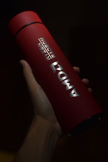 A red cylindrical tumbler featuring the AMD branding held in a hand. The lighting highlights the silver logo and text, creating a contrast against the dark background.