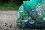 A green mesh bag filled with various empty cans and plastic bottles sits on a gravel surface. The background features some greenery, suggesting an outdoor setting.