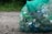 A green mesh bag filled with various empty cans and plastic bottles sits on a gravel surface. The background features some greenery, suggesting an outdoor setting.