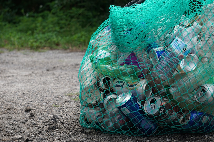 A green mesh bag filled with various empty cans and plastic bottles sits on a gravel surface. The background features some greenery, suggesting an outdoor setting.