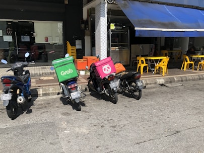 Multiple motorcycles are parked outside a small eatery, with two prominently displaying delivery boxes for delivery services. Yellow chairs and tables are set up under a blue awning next to the eatery.