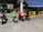 Multiple motorcycles are parked outside a small eatery, with two prominently displaying delivery boxes for delivery services. Yellow chairs and tables are set up under a blue awning next to the eatery.