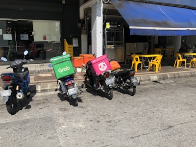 Multiple motorcycles are parked outside a small eatery, with two prominently displaying delivery boxes for delivery services. Yellow chairs and tables are set up under a blue awning next to the eatery.