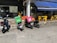 Multiple motorcycles are parked outside a small eatery, with two prominently displaying delivery boxes for delivery services. Yellow chairs and tables are set up under a blue awning next to the eatery.