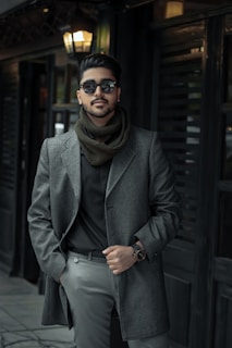 A fashionable man wearing a gray coat and sunglasses stands confidently against a dark, stylish background, with a warm light illuminating his face.