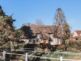 A charming cottage with a thatched roof is surrounded by lush greenery and tall trees. The building features small windows, and the front yard is decorated with hedges and flowers. A white fence and a path lead up to the entrance, creating a cozy and inviting atmosphere.