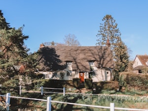 A charming cottage with a thatched roof is surrounded by lush greenery and tall trees. The building features small windows, and the front yard is decorated with hedges and flowers. A white fence and a path lead up to the entrance, creating a cozy and inviting atmosphere.