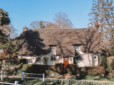 A charming thatched-roof cottage surrounded by well-manicured greenery. The cottage features small windows and a front door adorned with a wreath. Bunting is strung across the facade, adding a festive element. Mature trees frame the scene, providing a picturesque backdrop.