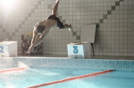 A male diver is captured in mid-air, diving into a swimming pool. The scene is set in an indoor pool area with tiled walls, and numbered starting blocks are visible. The water in the pool is calm, with lane ropes in place, indicating a competitive or training environment.