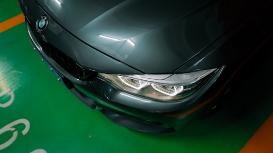 A dark-colored BMW car is parked in a green parking space. The focus is on the front end of the car, highlighting the headlights and the emblem on the hood. The lighting creates reflections on the paint, adding depth to the image.