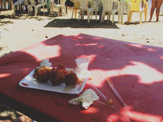 A red picnic table is shaded by overhead trees, casting natural patterns of light and shadow. On the table is a plastic plate with fried snacks, a cup, and some napkins. Plastic chairs and people are visible in the background, suggesting a relaxed outdoor setting.