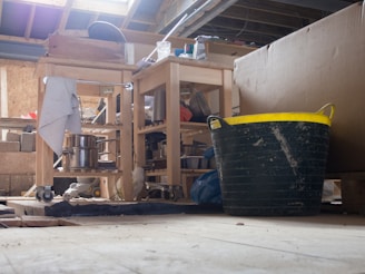 A cluttered workshop or storage area with wooden shelves on wheels holding various kitchen items, including metal pots and pans. A large black and yellow plastic tub is prominent in the foreground. There are bricks, scattered tools, and other miscellaneous objects in the background. The space appears dusty and has an unfinished ceiling with wooden beams.