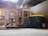 A cluttered workshop or storage area with wooden shelves on wheels holding various kitchen items, including metal pots and pans. A large black and yellow plastic tub is prominent in the foreground. There are bricks, scattered tools, and other miscellaneous objects in the background. The space appears dusty and has an unfinished ceiling with wooden beams.