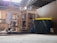 A cluttered workshop or storage area with wooden shelves on wheels holding various kitchen items, including metal pots and pans. A large black and yellow plastic tub is prominent in the foreground. There are bricks, scattered tools, and other miscellaneous objects in the background. The space appears dusty and has an unfinished ceiling with wooden beams.