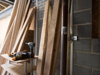 A collection of wooden planks leaning against a brick wall in a construction space. A power drill and other tools are placed on a makeshift wooden table nearby. The environment suggests an ongoing building or renovation project.