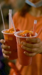 A person is holding two clear plastic cups filled with a red fruit smoothie topped with pieces of fruit and straw. The background is blurred, focusing attention on the drinks.