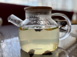 A clear glass teapot with a wooden lid, filled with light-colored liquid, possibly tea. The teapot's surface is slightly fogged, and it is sitting on a reflective surface. Inside the teapot, there are visible slices of fruit or herbs.