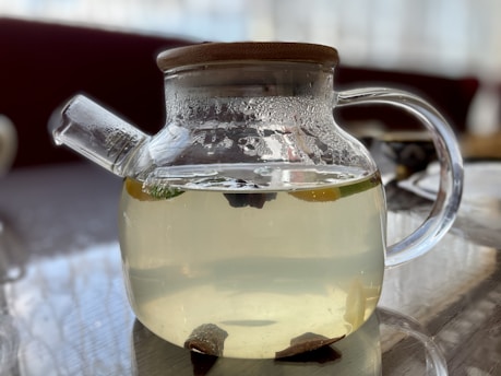 A clear glass teapot with a wooden lid, filled with light-colored liquid, possibly tea. The teapot's surface is slightly fogged, and it is sitting on a reflective surface. Inside the teapot, there are visible slices of fruit or herbs.