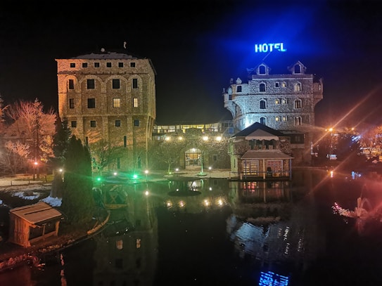 A nighttime scene features a large stone building with several windows, illuminated in warm lights. The building has a sign with the word 'HOTEL' glowing in blue. In the foreground, there is a body of water reflecting the lights and structures. Vegetation and a smaller wooden structure are visible on the left.