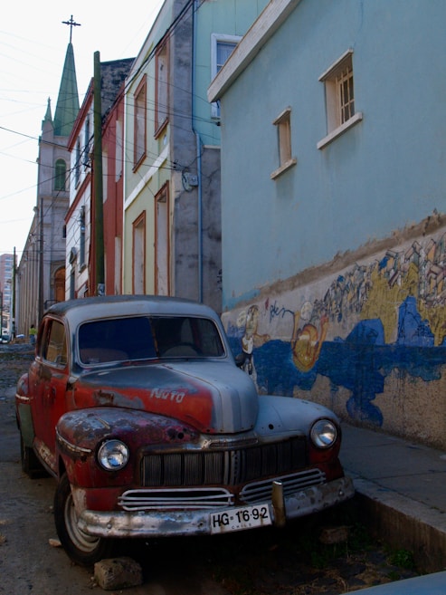 A vintage, weathered car with visible rust and peeling paint is parked along a narrow urban alleyway. The car's license plate indicates it is from Chile. To the left, a series of colorful, slightly worn buildings with a church steeple in the background create a classic street scene. On the right, a wall features a mural with various colors and designs.