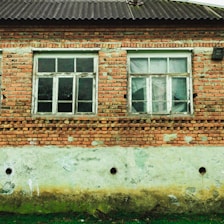 A brick house facade with two old, white-framed windows. The bricks are weathered and some mortar has come away, revealing patches of gray underneath. The lower part of the wall is a deteriorated concrete with signs of moisture damage, discoloration, and small holes. There is some green grass growing at the base of the wall.