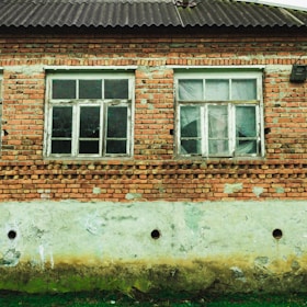 A brick house facade with two old, white-framed windows. The bricks are weathered and some mortar has come away, revealing patches of gray underneath. The lower part of the wall is a deteriorated concrete with signs of moisture damage, discoloration, and small holes. There is some green grass growing at the base of the wall.
