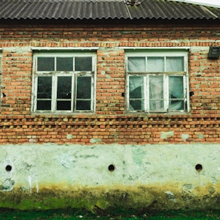 A brick house facade with two old, white-framed windows. The bricks are weathered and some mortar has come away, revealing patches of gray underneath. The lower part of the wall is a deteriorated concrete with signs of moisture damage, discoloration, and small holes. There is some green grass growing at the base of the wall.