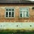 A brick house facade with two old, white-framed windows. The bricks are weathered and some mortar has come away, revealing patches of gray underneath. The lower part of the wall is a deteriorated concrete with signs of moisture damage, discoloration, and small holes. There is some green grass growing at the base of the wall.