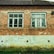 A brick house facade with two old, white-framed windows. The bricks are weathered and some mortar has come away, revealing patches of gray underneath. The lower part of the wall is a deteriorated concrete with signs of moisture damage, discoloration, and small holes. There is some green grass growing at the base of the wall.