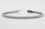 A delicate tennis bracelet featuring a series of equally sized, sparkling diamonds set in a row. The stones are mounted in a sleek metallic setting, forming an elegant and continuous line. The clasp is visible at one end, demonstrating the closure mechanism.