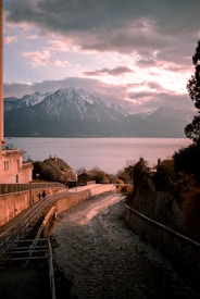 A serene landscape featuring a pathway beside a narrow water channel leading towards a tranquil lake with snow-capped mountains in the background. The sky is partly cloudy, illuminated by a warm sunset glow that casts a gentle light on the scene. Two figures are walking along the path, adding a sense of scale and tranquility to the composition.