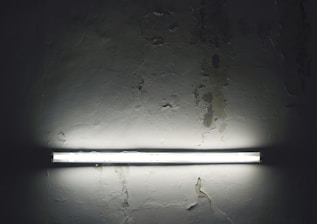 A fluorescent light fixture is mounted on a textured, worn concrete wall. The light is illuminating the immediate surrounding area, highlighting the cracks and rough patches on the surface. The ambience is dim and contrasts with the bright strip of light.