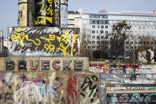 A stone structure with the word 'BRUECKE' prominently displayed, heavily adorned with colorful graffiti and street art. The background features a large building with the sign 'WIENER STÄDTISCHE' and other urban elements.