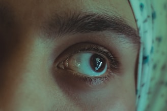 A close-up photo focusing on a person's eye, highlighting the details of the eyebrow and eyelashes. The skin tone is smooth, and there's a piece of patterned fabric partially visible on the right side.