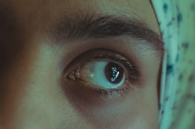 A close-up photo focusing on a person's eye, highlighting the details of the eyebrow and eyelashes. The skin tone is smooth, and there's a piece of patterned fabric partially visible on the right side.