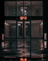A building entrance with a double door and a glowing sign displaying 'B-4' above it. The exterior walls feature decorative patterns, and an artistic light fixture with circular designs is visible through the upper windows. The wet ground reflects the scene, enhancing the moody ambiance.